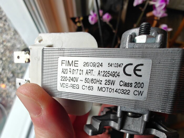 Närbild av en elektrisk komponent med märkning: FIME, 220-240V, 25W, och CE-märkning. Komponenten hålls i handen och har en typskylt.
