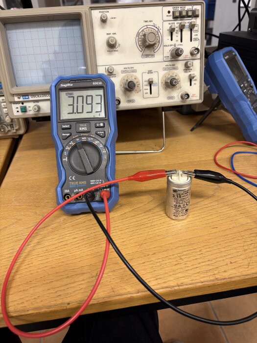 Multimeter som visar 3.093 V, ansluten till en kondensator på ett bord med gammal oscilloskoputrustning i bakgrunden.