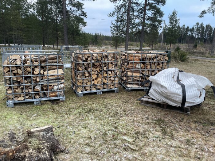 Fyrkantiga metallburar fulla med staplad ved står utomhus på en gräsbevuxen plats, bredvid en vit säck.