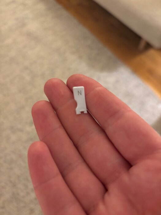 En bruten vit plastdel, märkt med "N", hålls i en hand mot en suddig bakgrund av ett vardagsrum.