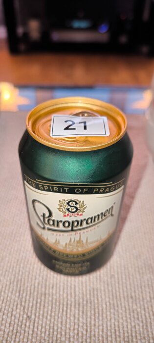 En burk Staropramen-öl med ett klistermärke med siffran 21 på locket, placerad på ett bord.