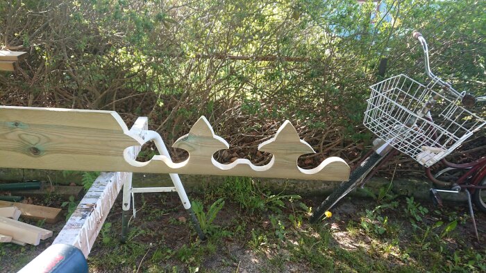 Dekorativ träsnittsdetalj på en bräda i en trädgård, liggandes på ett bockbord med cykel och grönska i bakgrunden.