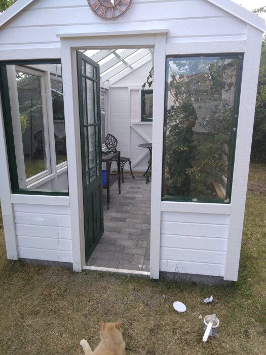 Växthus byggt av gamla fönster, öppet till dörr och en brun katt ligger framför. Inuti finns stolar och ett bord.