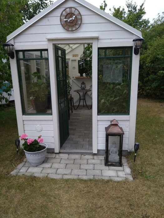 Växthus byggt av återvunna fönster, med en klocka på fasaden, en lykta med Buddha-statyn och blomkruka vid ingången, omgiven av grönska.