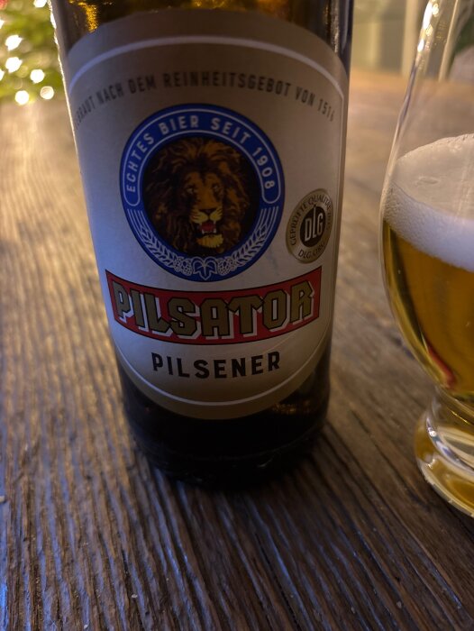 Flaska med Pilsator Pilsener på ett träbord, bredvid ett glas med delvis upphälld öl, etikett med lejonmotiv synlig.