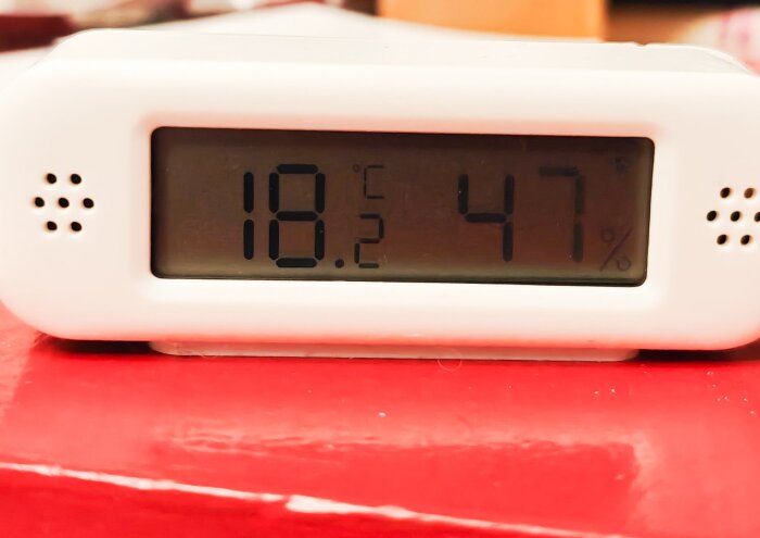 Digital termometer visar 18,2°C och 47% luftfuktighet, placerad på röd yta.