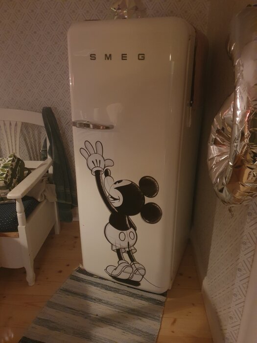 Ett vitt Smeg-kylskåp med en tecknad musfigur på, placerat i ett rum med mönstrad tapet och en stol bredvid.