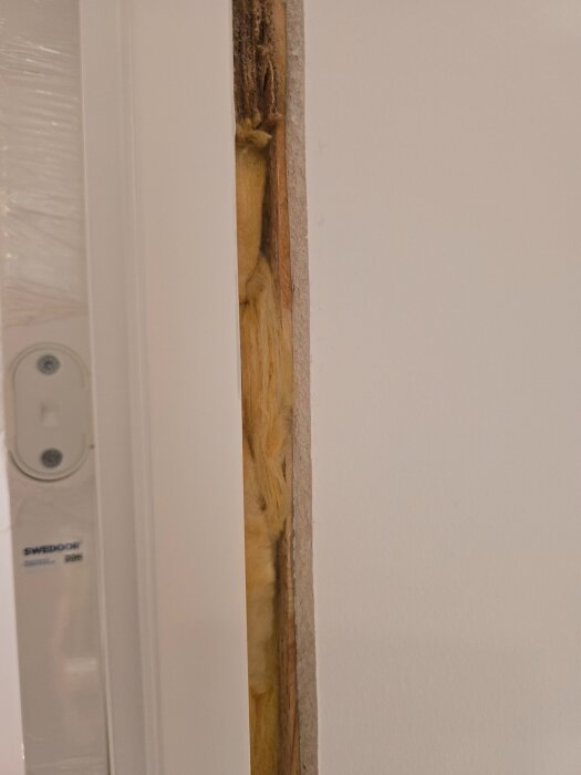 Närbild av en dörrkarm under installation, med glasull som isolering och skyddsfog för ljud- och brandskydd synlig mellan vägg och karm.