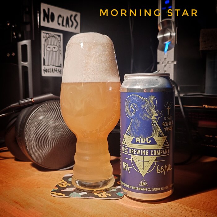 Ett glas öl med en skumkrona bredvid en burk från Apex Brewing Company, IPA 6.5%, på ett underlägg vid ett skrivbord.
