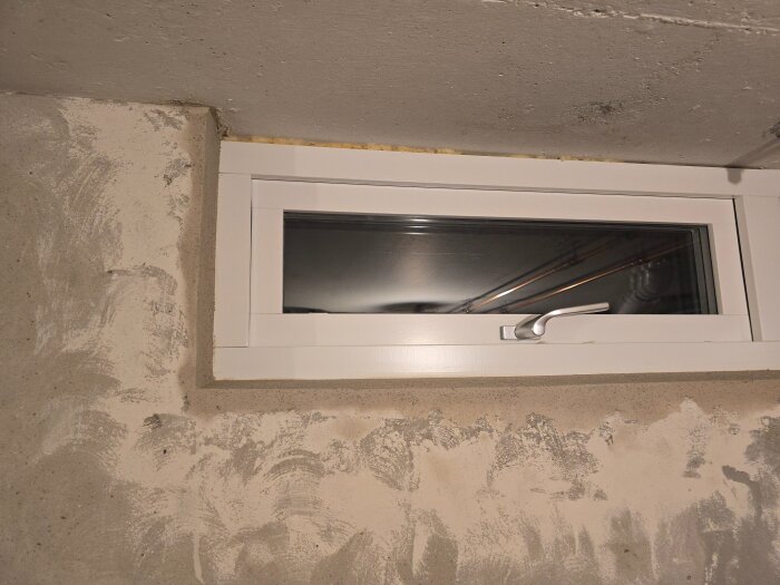 Vitt innanförliggande fönster med handtag i nyputsad grå betongvägg, synlig isolering ovanför fönstret.