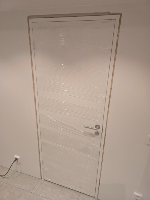 Vit innerdörr med plast runt, omgiven av ljusa väggar och ett trägolv, en övergång i arbetet med att installera dörrkarmar och ljudisolering.