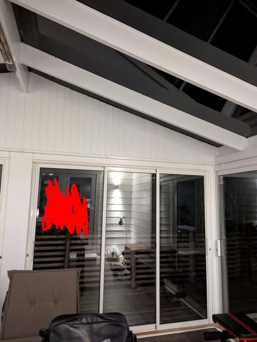 Inglasat uterum med vitmålade träbjälkar och skjutdörrar i glas. Ett bord syns genom glaset, rummet är delvis möblerat med fåtölj och lampa.