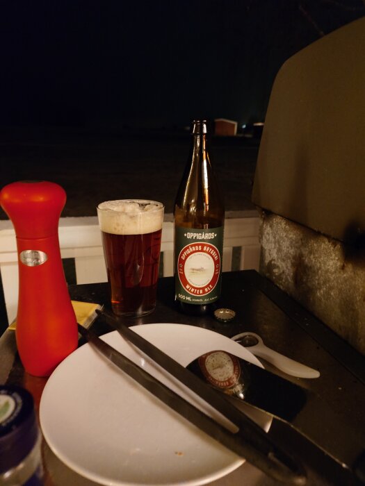 Flaska och glas med Oppigårds öl vid grillen, rödfärgad pepparkvarn och tallrik med verktyg ses i förgrunden.