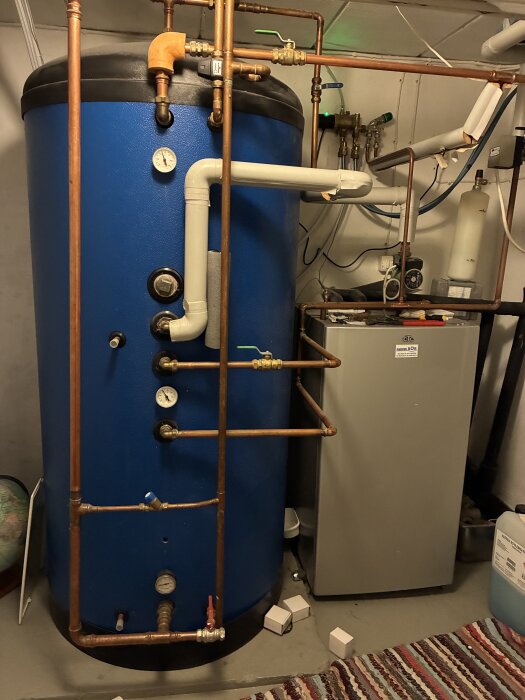 Ackumulatortank och rörsystem i ett värmesystem med rörkopplingar och mätare, fotad i ett pannrum med matta på golvet.