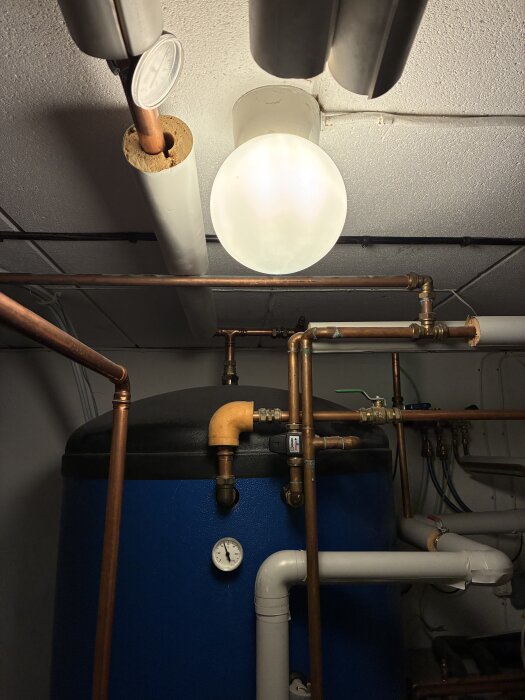 Ackumulatortank med kopparrör, termometer och isolerade rör i ett värmesystem, belyst rumstak i bakgrunden.