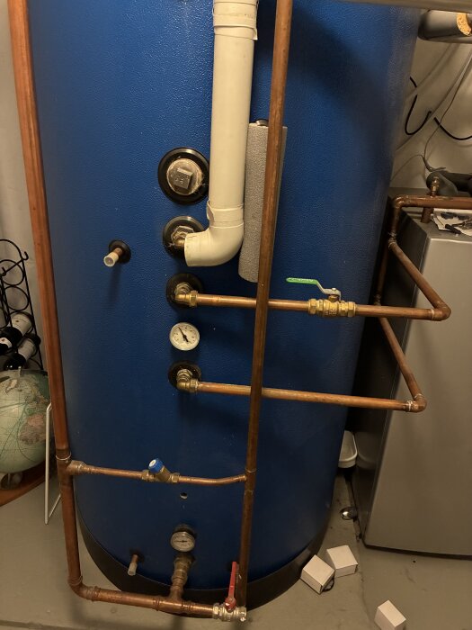 Blå ackumulatortank med kopparrör, ventiler och temperaturmätare i ett pannrum.
