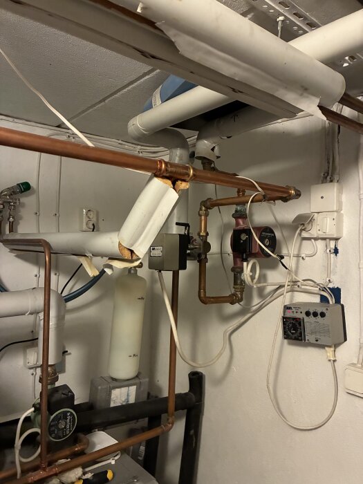 Uppvärmningssystem med kopparrör, cirkulationspump och elektronisk styrbox synliga på väggen i ett pannrum.