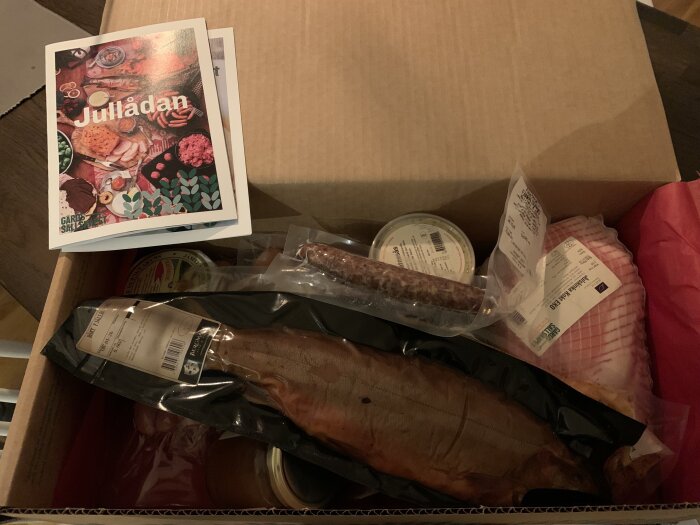 Julklappslåda med charkvaror, inklusive korv, rökt kött och inläggningar, tillsammans med broschyr som beskriver produkterna och deras ursprung.
