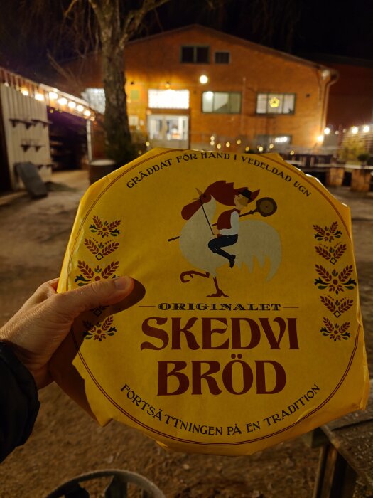 En hand håller ett paket Skedvi Bröd framför bageriet i St Skedvi, belyst på kvällen. Text på paketet: "Gräddat för hand i vedeldad ugn".