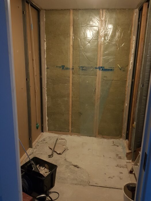 Ett rivet badrum med exponerade väggreglar och isolering, plaströr som sticker ut, verktyg och byggsopor på betonggolvet.