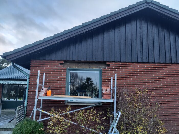 Tegelvägg med ett fönster under renovering, byggställning placerad framför, lövverk och himmel i bakgrunden.