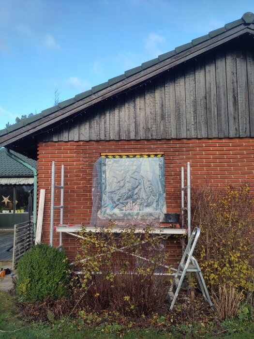 Fasad med fönster täckt av plast, stege och byggställning syns. Förbereder för murning, omgiven av buskar och trähusets tak i bakgrunden.