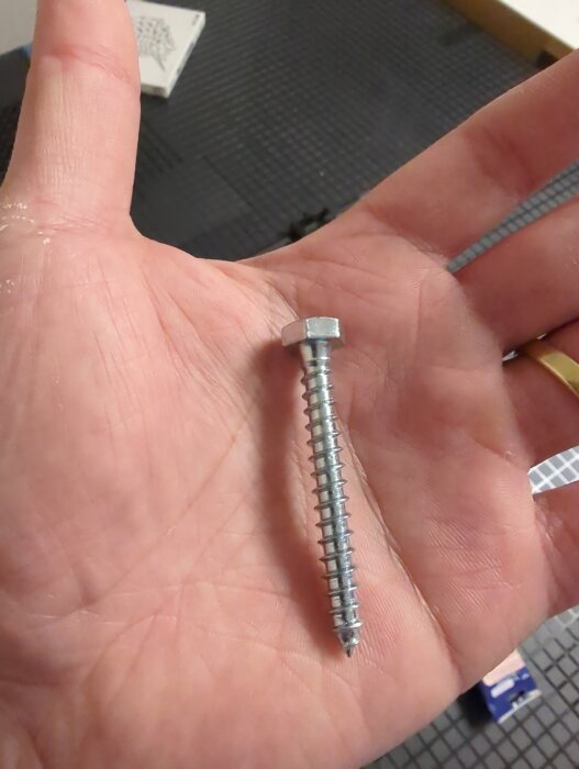 Hand håller i en metallskruv med hexagonalt huvud och gängor, avsedd för väggfäste, mot en bakgrund av verkstadsmatta.