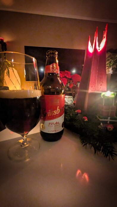 Mörk öl i glas och flaska bredvid röda ljus och juldekoration på bord.