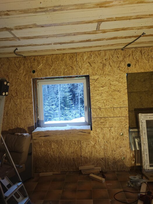 Ett nyinstallerat fönster i ett rum under renovering, med snö utanför, träpaneler på väggen och byggmaterial på golvet.