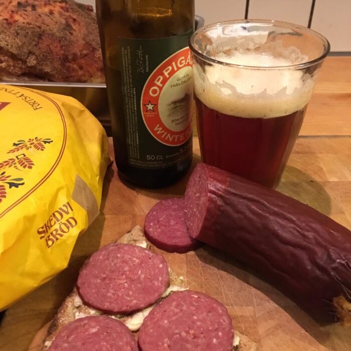 Skedvi bröd med skivad korv, en flaska och glas Oppigårds öl på ett träbord.