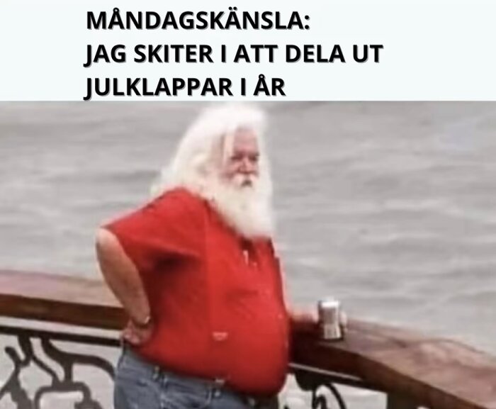 En äldre man med vitt skägg och röd t-shirt står vid ett räcke och håller i en dryck. Text ovan: "Måndagskänsla: Jag skiter i att dela ut julklappar i år.