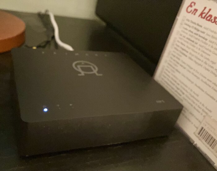 HiFi-strömmare på ett bord blinkar med en blå lampa, omgiven av sladdar och en bok eller manual bredvid.