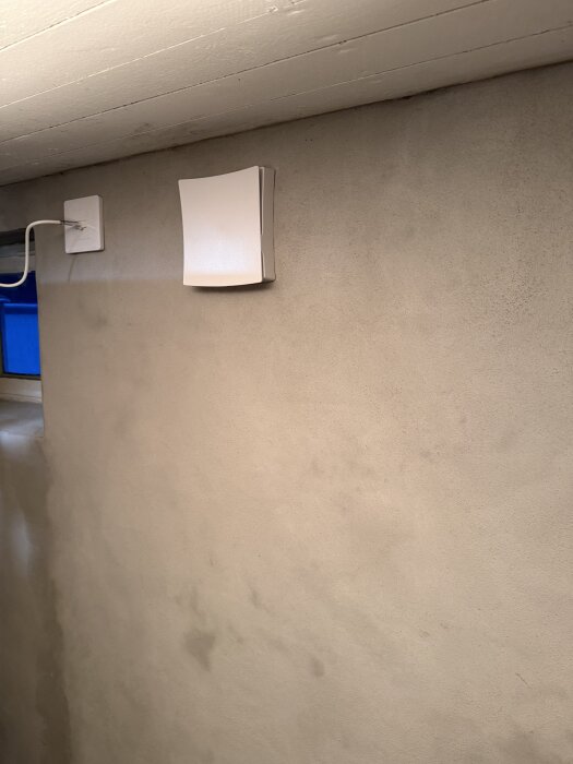 Ventilationsfläkt installerad på en putsad vägg i ett rum med en liten, blåmålad del av ett fönster synlig i bakgrunden.