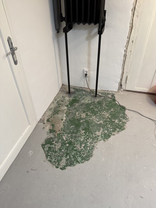 Slitet golv med flagande grön färg i hörn av rum med vit vägg och svart element över en eluttag.