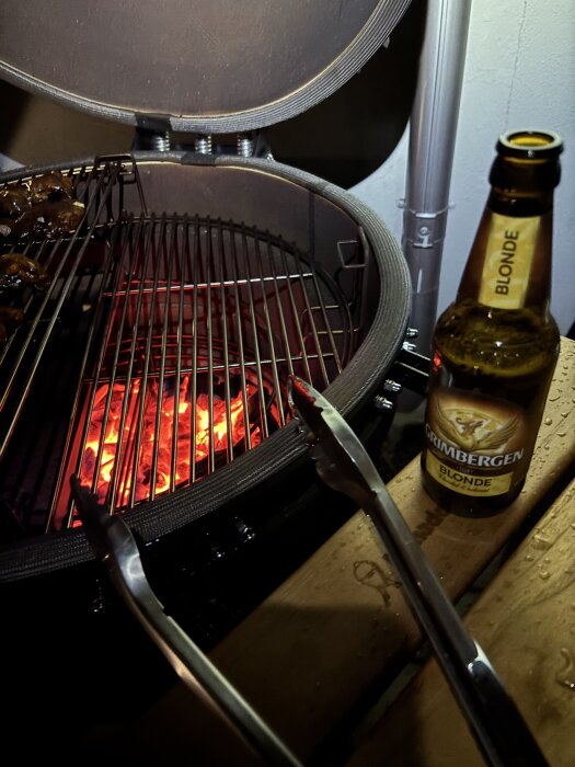 Grill med heta kol, grilltång och en öppnad flaska öl på ett träbord.