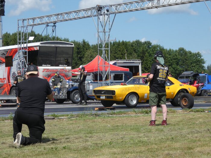 En gul sportbil och en man i svarta kläder vid en racerbana, omgivna av åskådare och bilar, under en solig dag med klart väder.