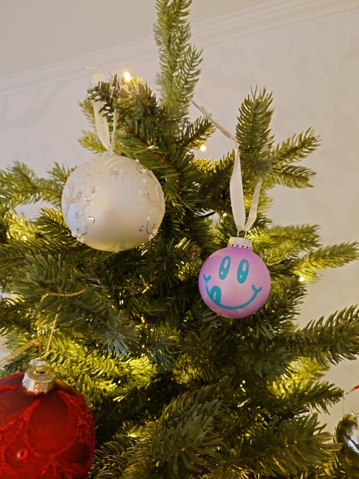 Julgransdekorationer med en vit kula och en lila kula med ansikte hänger i granen.