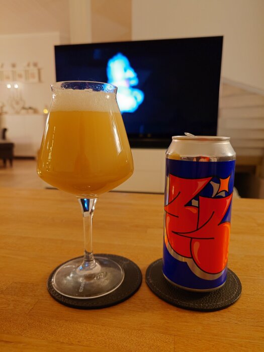 Ett glas öl och en ölburk står på ett träbord med en TV i bakgrunden som visar ett oskarpt ljusmönster.