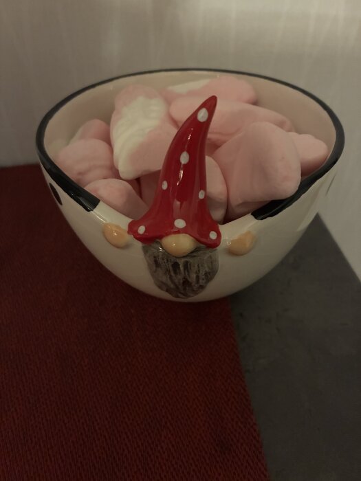 Skål med en tomtefigur fylld med rosa och vita godishjärtan, placerad på en mörkröd duk.