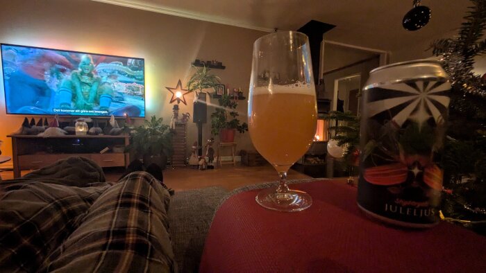 Person i pyjamas tittar på julfilm med julöl i glas framför brasa, dekorerat vardagsrum och tv-skärm visar film.