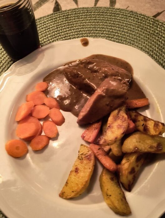Tallrik med kyckling i mörk sås, ugnsrostade potatisar, morotsskivor och ett glas julmust bredvid på en grön bordstablett.