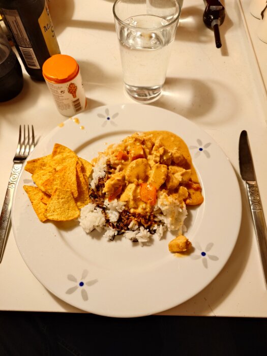 Tallrik med kycklingrätt, sås, vitt ris, nachochips och ett glas vatten. Sojaflaska i bakgrunden på bordet.