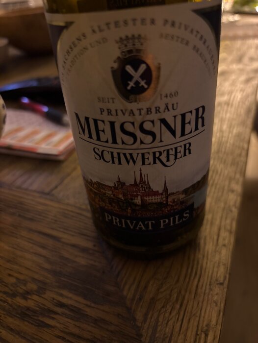 Flaska med etikett från Meissner Schwerter Privat Pils bryggeri, med text och en illustration av en stadssilhuett i bakgrunden, på ett träbord.