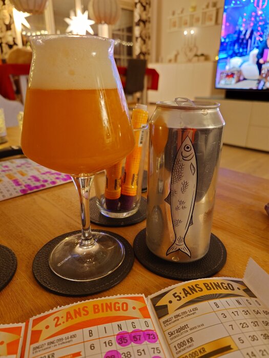 Ett glas öl och en burk med fiskdesign på ett bord, omgivna av bingobrickor och en tänd TV i bakgrunden.