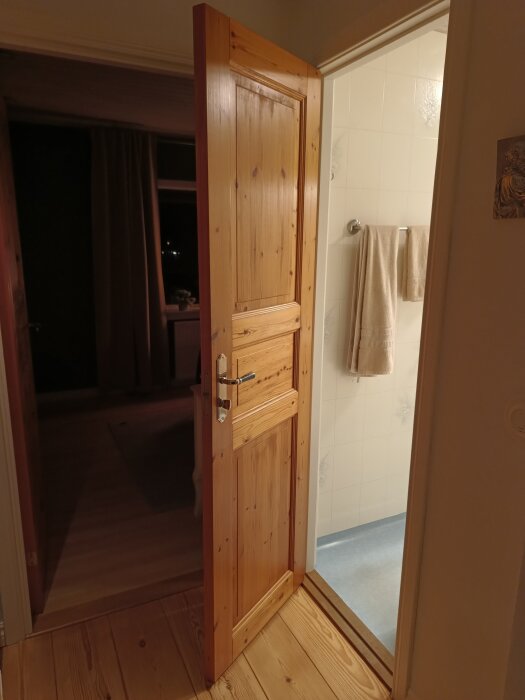 Trädörr till badrum som verkar ha varit i bruk i 30-40 år, öppnad mot ett rum med handdukshängare och klinkers på golvet.