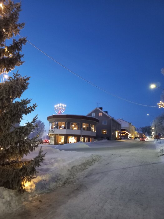 En snötäckt gata i skymningen med julbelysning, ett tvåvåningshus och en upplyst gran.