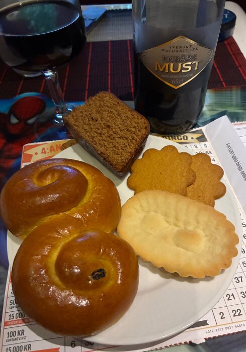 Tallrik med lussekatter, mjuk pepparkaka, pepparkakor och kex, samt ett glas och flaska med enbärsmust på bordet.