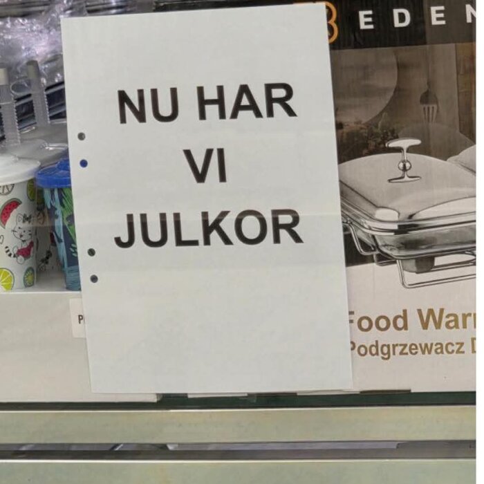 Skylt med texten "Nu har vi julkor", omgiven av plastmuggar och en förpackning med en matvarmhållare i bakgrunden.