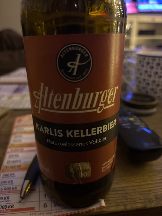 Flaska Altenburger Karlis Kellerbier på Bingolottokupong med penna och fjärrkontroll i bakgrunden.