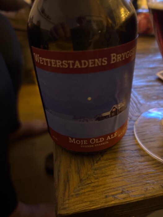 Flaska av Wetterstadens Bryggeri Moje Old Ale på ett träbord bredvid ett halvt fyllt glas, etikett med vinterlandskap och text.